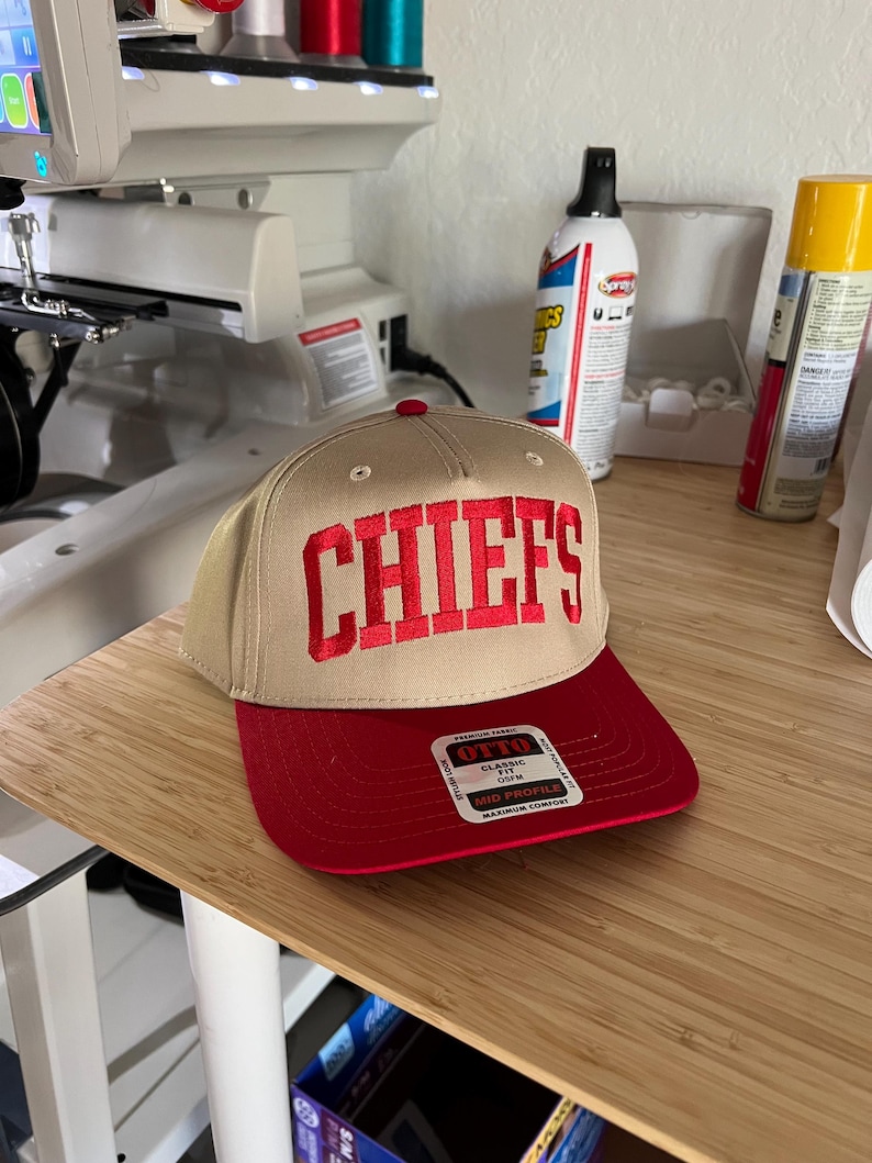 May include: A tan baseball cap with a red brim and the word "CHIEFS" embroidered in red on the front. The cap has a black and white label that says "OTTO CLASSIC FIT OSFM MID PROFILE MAXIMUM COMFORT".