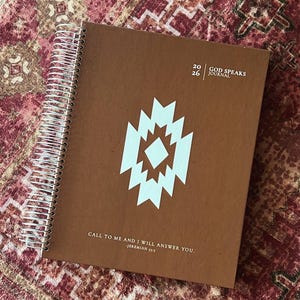 May include: A brown spiral-bound journal with a white geometric diamond-shaped emblem in the center. The cover features the text 2026 GOD SPEAKS JOURNAL and the quote CALL TO ME AND I WILL ANSWER YOU. -JEREMIAH 33:3 printed in white ink.