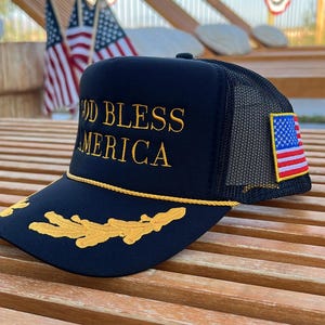 May include: A navy blue trucker hat with the text "GOD BLESS AMERICA" embroidered in gold. The hat features a mesh back, an American flag patch, and gold decorative accents. The hat is sitting on a wooden surface.