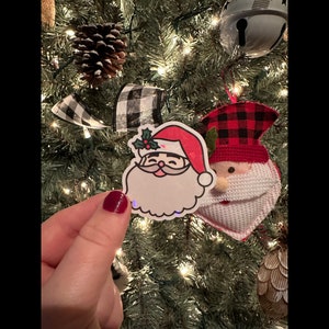 May include: A red and white Christmas ornament with a cartoon Santa Claus face. The ornament is hanging on a Christmas tree with green needles and white lights.