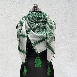 May include: A white and green patterned scarf with green tassels, displayed on a black mannequin. The scarf features a checkered design in green and white, along with green stripes and tassels adorning the edges.