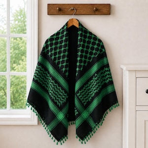 Peut inclure: Un keffieh noir et vert orné d'un motif traditionnel à carreaux et chevrons. Ce tissu de forme carrée est suspendu à un cintre en bois et présente de petites franges vertes assorties le long des bords inférieurs du vêtement.