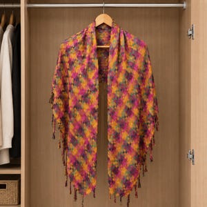 Puede incluir: Una bufanda rectangular con un vibrante patrón de diamantes multicolor en tonos magenta, amarillo mostaza y morado oscuro. La tela tiene una textura tejida y está terminada con flecos decorativos a lo largo de los bordes inferiores.