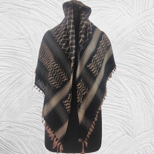 Puede incluir: Un pañuelo cuadrado con un patrón geométrico en negro y bronceado con un diseño de cuadros tradicional. La tela muestra un motivo de zigzag repetitivo y bandas anchas sólidas, terminado con flecos anudados decorativos en los bordes.