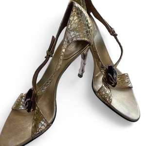 Peut inclure: Une paire d'escarpins slingback dorés avec une finition texturée effet peau de serpent. Chaque chaussure présente un bout pointu, un talon aiguille fin et un ornement sombre et brillant sur la bride en T. Inclut des brides de cheville réglables.
