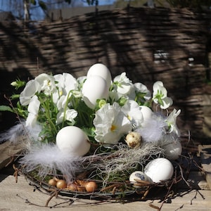 Könnte beinhalten: Ein österliches Arrangement mit weißen Eiern, weißen Blumen und kleinen, gefleckten Wachteleiern. Die Eier sind zwischen den Blumen und dem Grün angeordnet, mit zarten weißen Federn. Das Arrangement befindet sich in einem rustikalen, geflochtenen Korb.