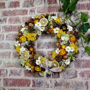 Könnte beinhalten: Ein runder Kranz aus getrockneten gelben Blumen, braunen Tannenzapfen, ganzen Haselnüssen und getrockneten Zitronenscheiben. Das Arrangement enthält kleine weiße Blüten und grüne Blätter, abgerundet durch ein braun-weiß kariertes Stoffband.