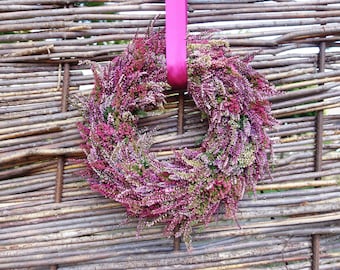 Könnte beinhalten: Ein kreisförmiger Kranz aus dichten Büscheln leuchtend magentafarbener, violetter und weißer Heidekrautblüten. Das Blumenarrangement ist an einem glatten, leuchtend pinkfarbenen Satinband befestigt, das durch die Mitte des Kranzes verläuft.
