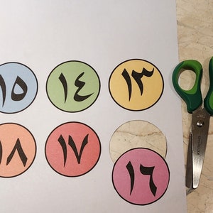 May include: A set of seven colourful paper circles with Arabic numerals 10 through 17 printed in black. The circles are arranged in a grid pattern on a white background. A pair of green handled scissors is on the right side of the image.