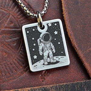 May include: Silver-toned astronaut pendant necklace. The square pendant features a detailed engraving of an astronaut on a lunar surface with stars in the background. The necklace has a chain link design and is displayed on a brown leather surface.