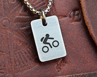 May include: A silver-colored pendant necklace featuring a rectangular tag with a black bicycle icon. The chain is a small, linked design. The pendant hangs against a brown leather background.