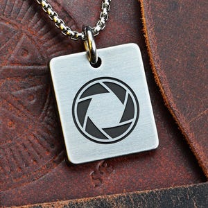 Op de afbeelding: Een zilverkleurige vierkante hangerketting met een zwart diafragma-symbool. De hanger hangt aan een zilveren ketting. De ketting wordt gepresenteerd op een bruin leren oppervlak. Het woord "FRONT" is zichtbaar linksboven.