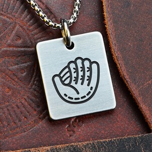 Puede incluir: Collar con colgante cuadrado plateado con diseño de guante de béisbol negro. El colgante cuelga de una cadena plateada. La palabra "FRONT" es visible en la esquina superior izquierda.