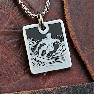 May include: Silver-tone pendant necklace. The rectangular pendant has a black top section and a surfer graphic. The chain necklace is visible against a brown leather background. The pendant is a stylish accessory.