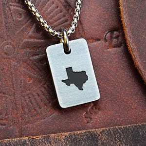 May include: A silver-colored rectangular pendant necklace featuring a black silhouette of the state of Texas. The pendant hangs from a silver chain. The background is a brown leather surface.