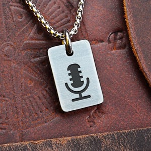 Könnte beinhalten: Eine silberfarbene Halskette mit Anhänger, die ein rechteckiges Schild mit einer schwarzen Mikrofon-Grafik zeigt. Die Halskette hat eine Kettenglieder-Optik und wird auf einem braunen Lederhintergrund präsentiert. Das Wort "FRONT" ist oben sichtbar.