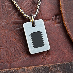 May include: A silver-coloured dog tag pendant featuring a black integrated circuit design, suspended from a matching chain. The pendant is rectangular with rounded corners. The background is a textured brown leather surface.
