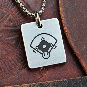 Puede incluir: Un collar con colgante cuadrado plateado con un diseño de campo de béisbol. El colgante cuelga de una cadena plateada. El collar se muestra sobre una superficie de cuero marrón. La palabra "FRONT" es visible en la esquina superior izquierda.