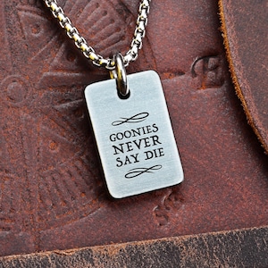 Könnte beinhalten: Halskette mit silberfarbenem Anhänger in Form einer Hundemarke mit dem Text "GOONIES NEVER SAY DIE". Der Anhänger hängt an einer silbernen Kette. Der Hintergrund ist eine braune Lederoberfläche.