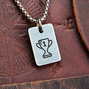 Puede incluir: Collar con colgante rectangular plateado con un contorno negro de un trofeo y el número 1. El colgante cuelga de una cadena plateada. El fondo es una superficie de cuero marrón.