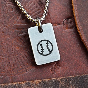 Puede incluir: Un collar con colgante rectangular plateado con un diseño de béisbol negro. El colgante cuelga de una cadena plateada. El fondo es una superficie de cuero marrón.
