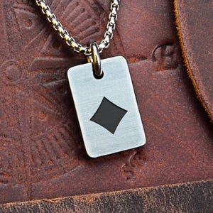 May include: A silver-colored dog tag pendant with a black diamond symbol, suspended from a silver chain. The tag is rectangular with rounded corners. The background is a brown leather surface.