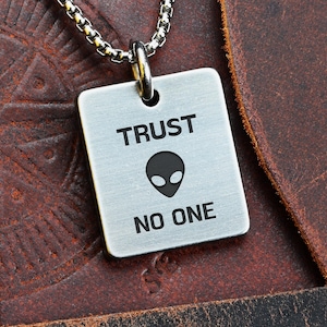 May include: A silver-colored square pendant necklace with the words "TRUST NO ONE" and an alien head graphic. The pendant hangs from a silver chain. The background is a textured brown leather surface.
