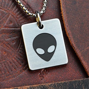 Può includere: Una collana con pendente quadrato color argento con un design a testa di alieno nero. Il pendente è appeso a una catena d'argento. La parola "FRONT" è visibile in alto a sinistra. Lo sfondo è una superficie in pelle marrone.