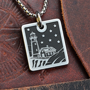 Puede incluir: Collar con colgante plateado que presenta un diseño de faro. El colgante rectangular tiene un fondo negro con estrellas blancas y una ilustración de faro. El collar se muestra sobre una superficie de cuero marrón.