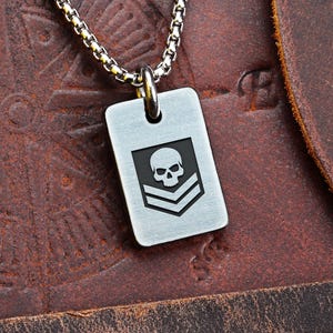 Puede incluir: Un colgante de placa de identificación plateado con un diseño de calavera y galones, suspendido de una cadena. El colgante es rectangular y presenta un gráfico detallado de calavera sobre dos galones. La cadena es de diseño pequeño y enlazado.