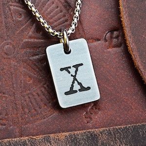 Può includere: Una collana con pendente color argento con un'etichetta rettangolare incisa con la lettera "X" in nero. Il pendente è appeso a una catena. Lo sfondo è una superficie in pelle marrone strutturata.