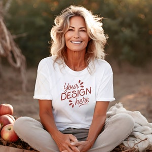 以下が含まれることがあります： 白いTシャツに、茶色の文字で「Your Design here」と書かれています。シャツは、屋外に座っている人が着ています。