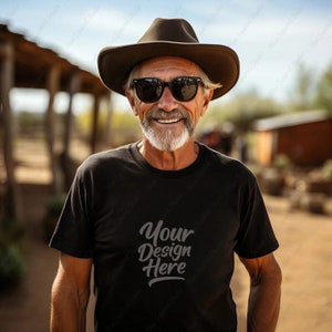Op de afbeelding: Een man draagt een zwart T-shirt met de tekst "Your Design Here" erop gedrukt. Hij draagt een bruine cowboyhoed en een zonnebril.