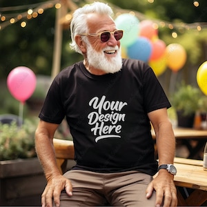 Könnte beinhalten: Ein Mann trägt ein schwarzes T-Shirt mit dem Text "Your Design Here" in weiß gedruckt. Er sitzt im Freien mit einem entspannten Gesichtsausdruck und trägt eine Sonnenbrille.