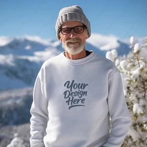 Puede incluir: Una sudadera blanca con el texto "Your Design Here" en una fuente cursiva gris. La sudadera la lleva puesta una persona con una boina gris y gafas, de pie frente a un fondo de montaña nevado.