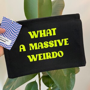 Könnte beinhalten: Schwarze Canvas-Kosmetiktasche mit dem Text "WHAT A MASSIVE WEIRDO" in leuchtendem Gelb.