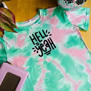 Könnte beinhalten: Ein grün-pinkes Batik-T-Shirt mit dem Text "HELL YEAH" in schwarzen Buchstaben.