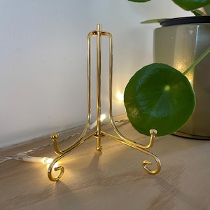 Puede incluir: Un soporte de exhibición de metal dorado con una parte superior redonda y tres patas. El soporte tiene un diseño decorativo de desplazamiento en las patas.