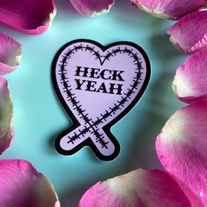 May include: A pink heart-shaped sticker with black stitching around the edges. The sticker says "HECK YEAH" in black letters.