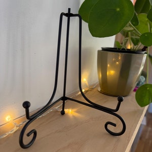 Könnte beinhalten: Ein schwarzer Metall-Ausstellungsständer mit einem dreibeinigen Sockel und einer geschwungenen Oberseite. Der Ständer ist für eine Platte oder ein Kunstwerk konzipiert. Der Ständer steht auf einem Holzregal, mit einer Pflanze in einem Topf im Hintergrund.