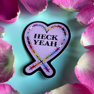 May include: A pink heart-shaped sticker with a black outline and colorful stitching around the edges. The sticker says "HECK YEAH" in black letters.