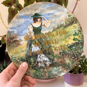 Op de afbeelding: Decoratieve bord met een schilderij van een vrouw in een groene jurk en hoed, tegen een landschap met een bewolkte hemel. Gouden tekst luidt "SEARCHING FOR MY FINAL FUCK TO GIVE". Het bord heeft een gouden rand.