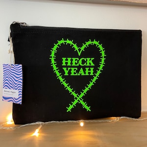 May include: Black canvas zippered pouch with a green heart design and the text "HECK YEAH" inside the heart.