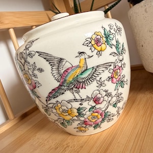 Op de afbeelding: Een crèmekleurige keramische pot met deksel, voorzien van een handgeschilderd ontwerp van een vogel met veelkleurige vleugels op een tak, omringd door gele en roze bloemen met groene bladeren, uitgevoerd in een gedetailleerde, fijne illustratiestijl.