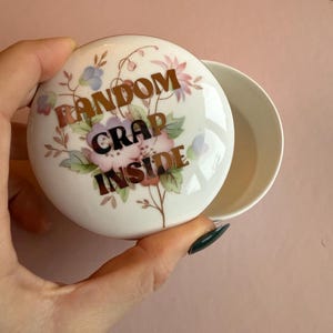 May include: A white ceramic trinket box with a lid featuring floral designs and the text "RANDOM CRAP INSIDE" in gold lettering. The box is open, revealing its interior.