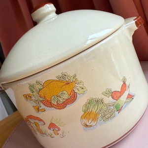 以下が含まれることがあります： 蓋付きの白い陶器製のキャセロール。キャセロールは茶色の縁取りと、野菜とキノコのカラフルなデザインが施されています。