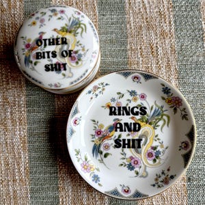 May include: A white ceramic trinket box and dish set with floral and bird designs. The box lid reads "OTHER BITS OF SHIT" and the dish reads "RINGS AND SHIT". Both pieces have gold trim.