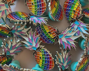 May include: A collection of enamel pins shaped like smiling pineapples. The pineapples feature a rainbow color scheme, with a grid pattern on the body and a silver outline. The pineapple tops are white, pink, and blue. The pins have a light blue backing.