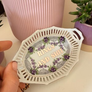 Peut inclure: Petit plat à bijoux en céramique blanche avec un bord doré et un design ajouré. Le centre porte l'inscription dorée "RINGS AND SHIT", entourée de motifs floraux violets. Une main tient le plat.