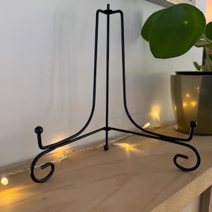 Puede incluir: Un caballete de metal negro con un diseño decorativo de desplazamiento en la base. El caballete está hecho de metal delgado y tiene un diseño simple y elegante.